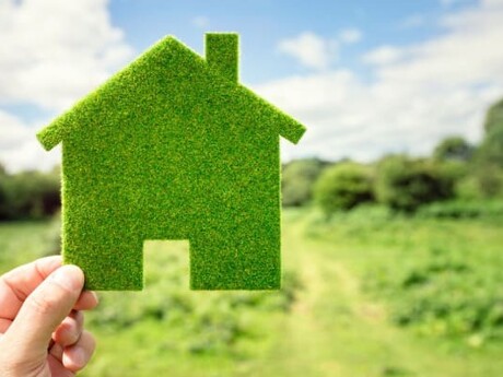 Eine Hand hält ein Hausmodell aus Gras vor einer Wiese hoch