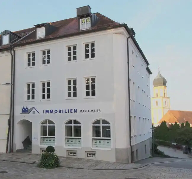 Weißes Gebäude "Immobilien Maria Maier", Kirchturm im Hintergrund.