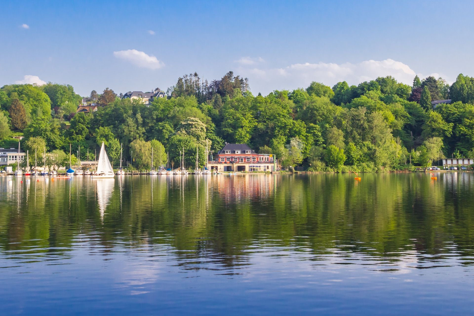 Baldeney-See in Essen mit Segelboot, Bootshaus und grünen Bäumen auf ruhigem Wasser.