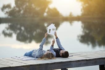Paar hält Modellhaus auf Steg am See bei Sonnenuntergang.