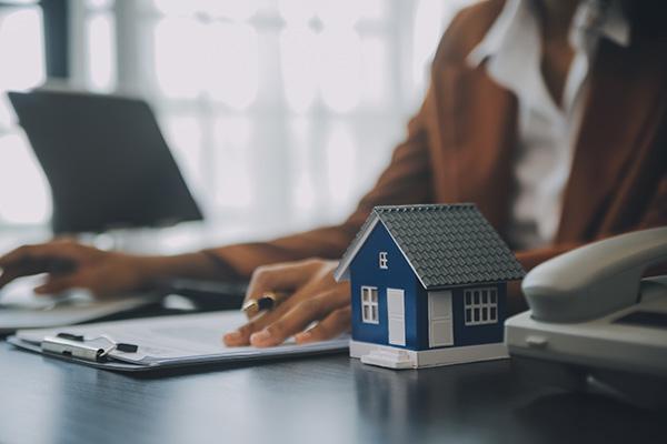 Person an Schreibtisch mit Klemmbrett, Laptop und Modellhaus.