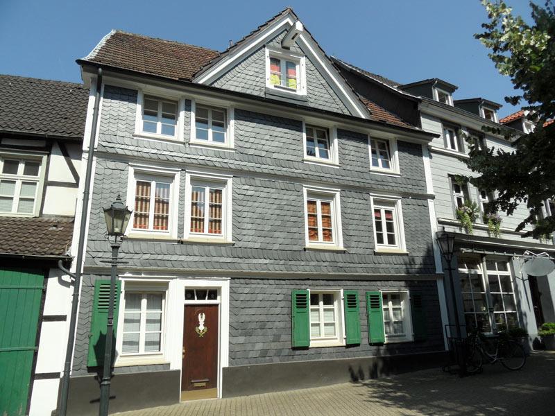 Graues Haus mit grünen Fensterläden, brauner Tür und Fenstern im Sonnenlicht.