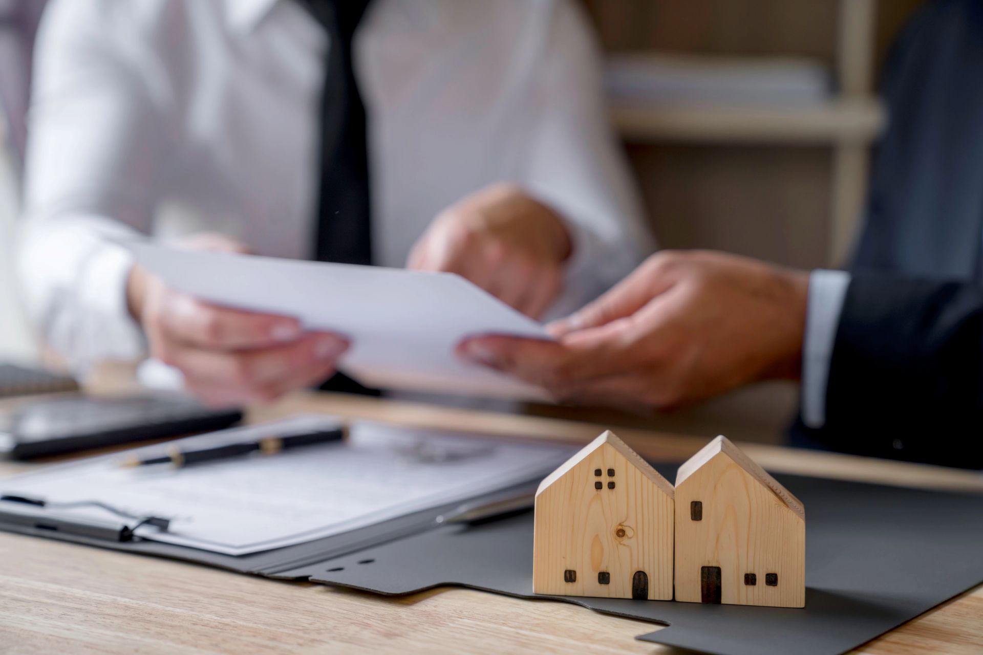 Zwei Personen prüfen Dokumente am Tisch, kleine Holzhäuser vorn.