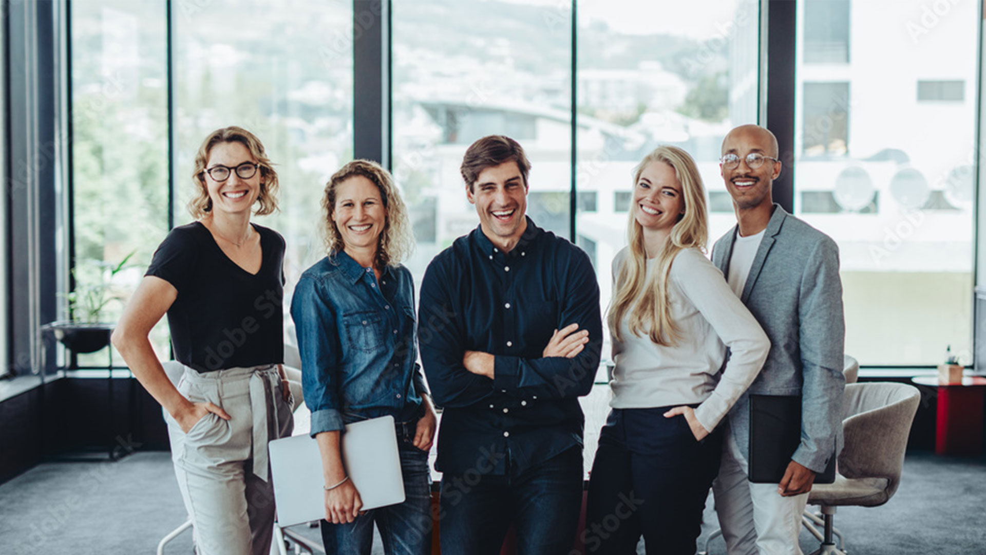 Fünf lächelnde Fachkräfte stehen im modernen Büro vor Fensterfront.
