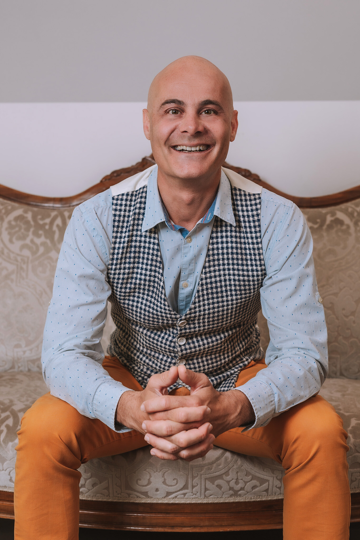 Uomo calvo in camicia blu, gilet e pantaloni arancioni su un divano decorato.