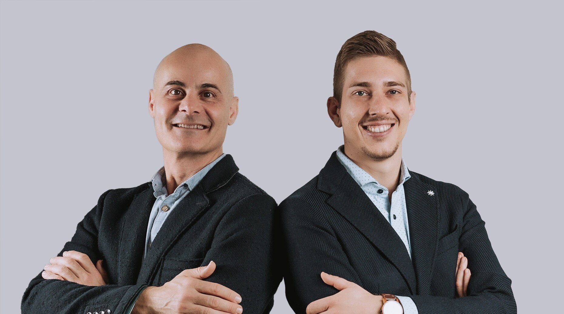 Zwei Männer in dunklen Blazern, Arme verschränkt, vor grauem Hintergrund.