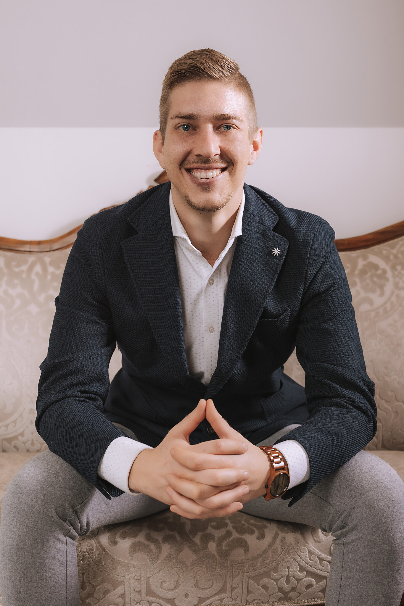 Un uomo sorridente in blazer siede sul divano a motivi, con le mani giunte.