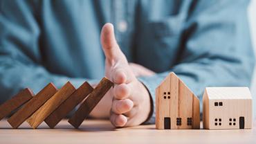 Eine Person hält fallende Dominosteine vor zwei Hausmodellen auf