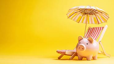 Ein Sparschwein steht vor einer Strandliege mit Sonnenschirm