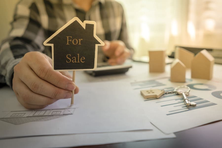 Eine Person am Schreibtisch hält ein "For Sale"-Schild in der Hand