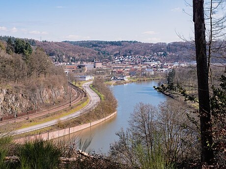 Fluss und Bahn neben kleinem Ort, umgeben von Hügeln und Bäumen.