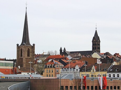 Stadtbild mit Kirchtürmen und roten Dächern unter bewölktem Himmel.