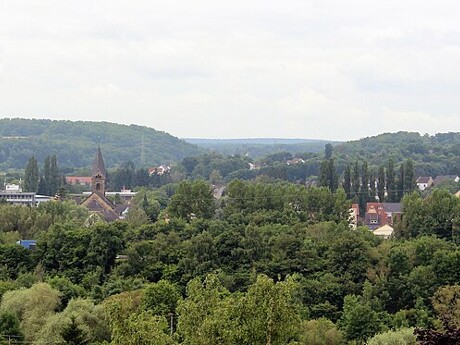 Kleinstadt mit Kirchturm, Bäumen, Hügeln und Wolken.
