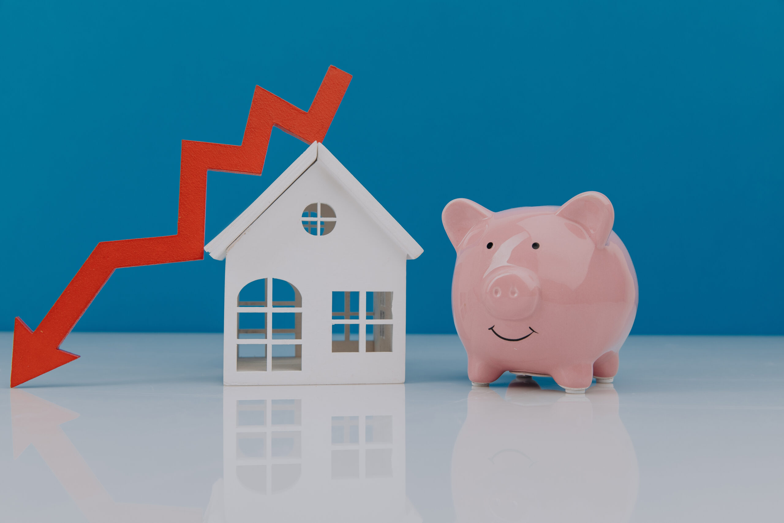 Sparschwein neben Modellhaus und sinkenden Pfeil