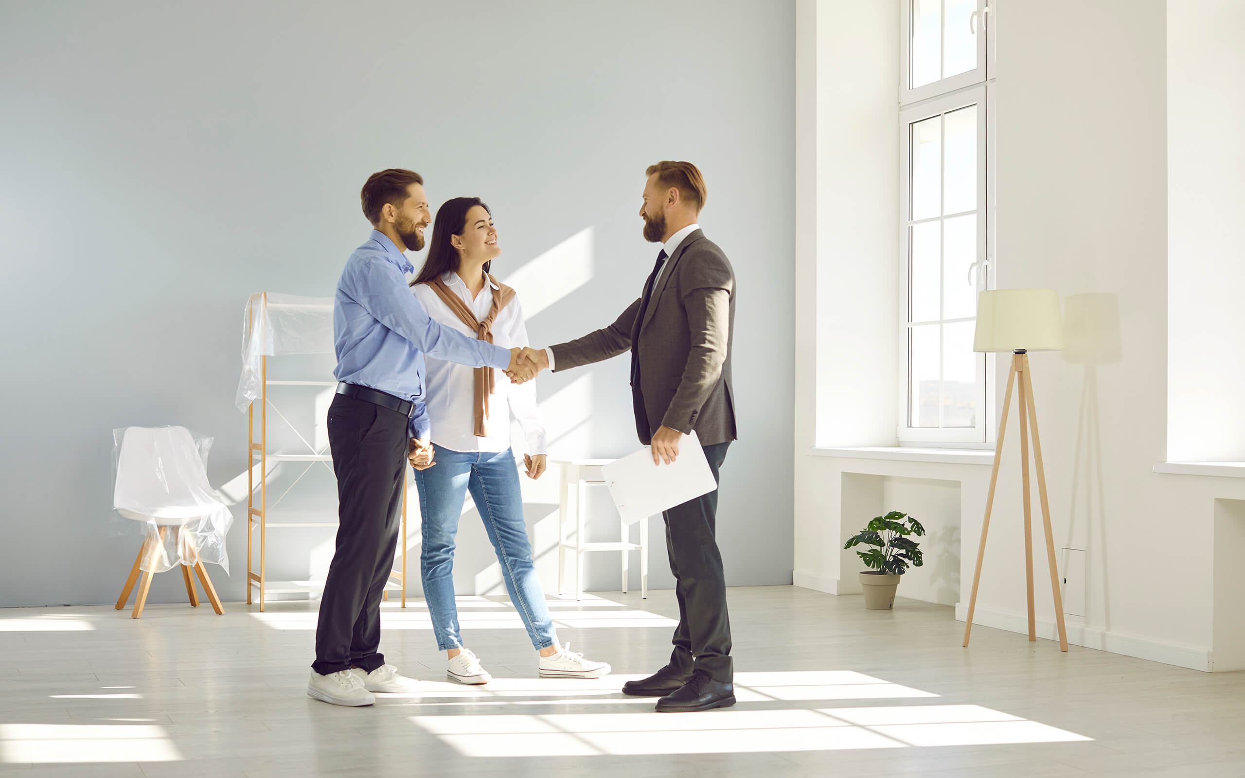 Paar und Makler geben sich in hellem, leerem Raum die Hand.