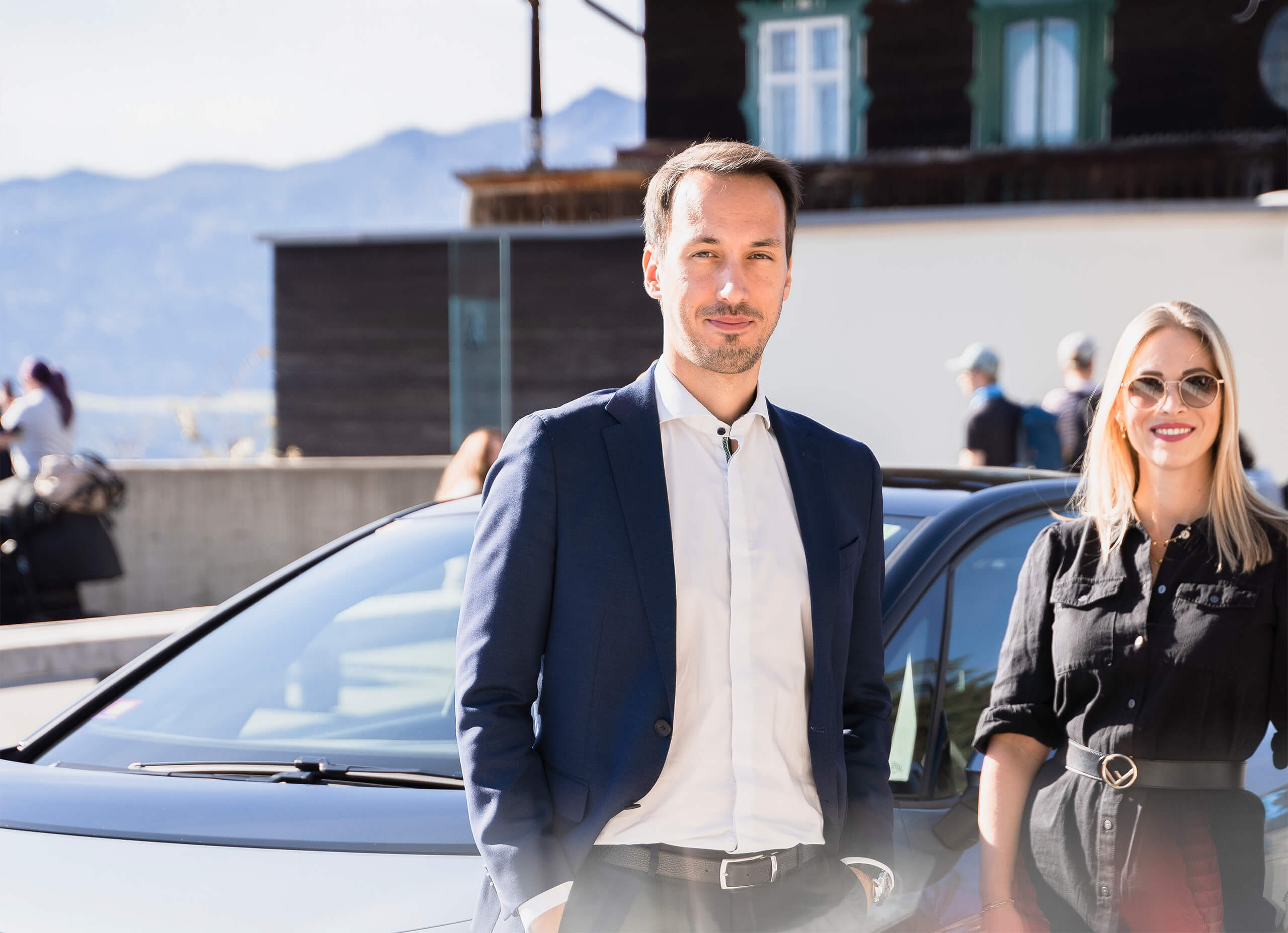 Mann im blauen Anzug und Frau mit Sonnenbrille lächeln vor Auto.
