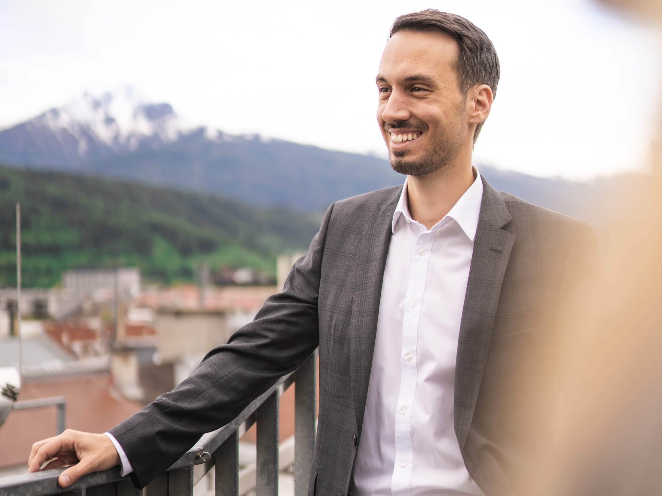 Lächelnder Mann im Anzug auf Balkon mit Bergen im Hintergrund.