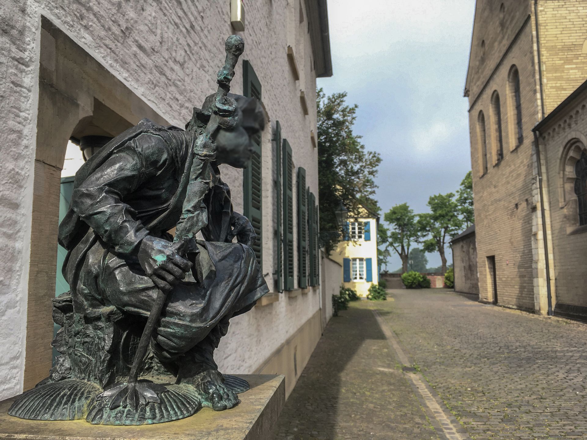 Bronzestatue einer vermummten Figur mit Stab auf einer Kopfsteinpflasterstraße.