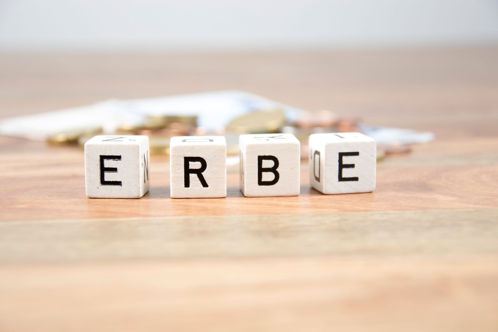 Würfel mit "ERBE" vor unscharfen Münzen und Dokumenten auf Holz.