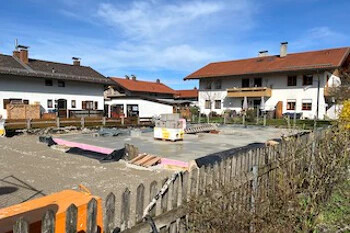 Baustelle mit Fundament, rote Dächer, Holzzaun im Vordergrund.