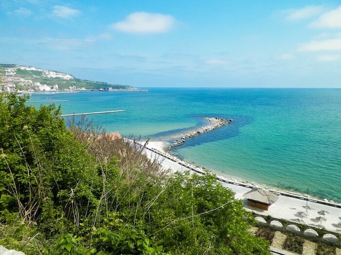 Blaues Meer, Sandstrand, grüne Büsche und helle Wolken am Himmel.