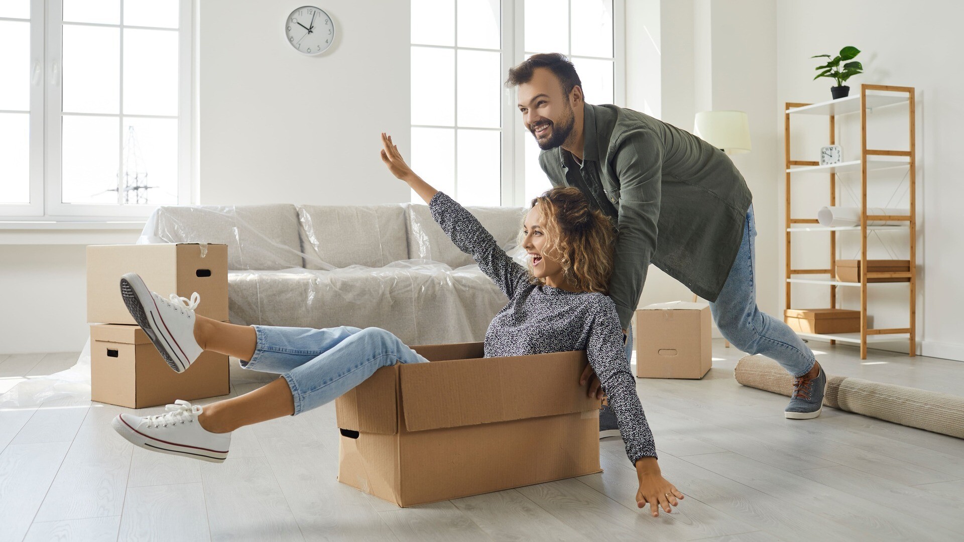 Ein Mann schiebt eine Frau, die in einem Umzugskarton sitzt, durch ihre neue Wohnung.