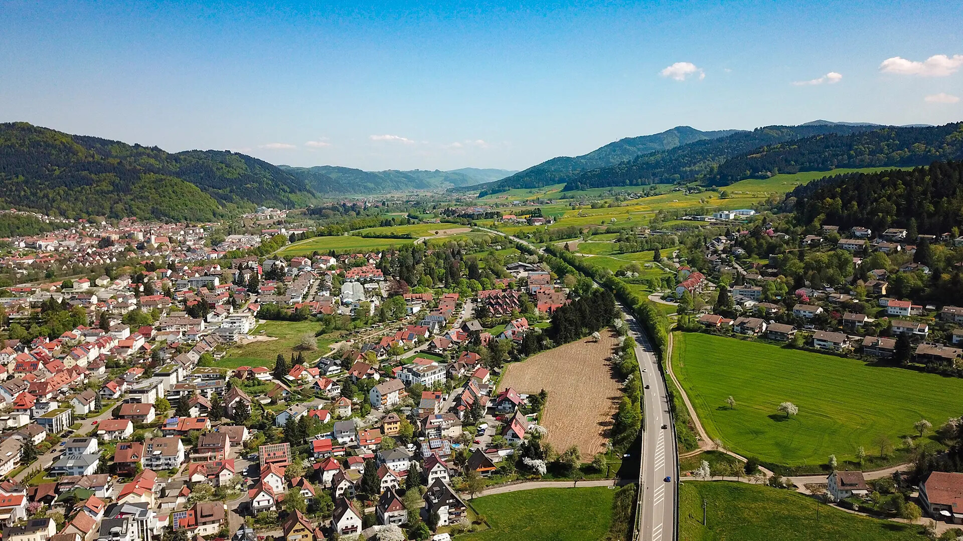 Kleinstadt mit roten Dächern, Feldern und Hügeln bei blauem Himmel.