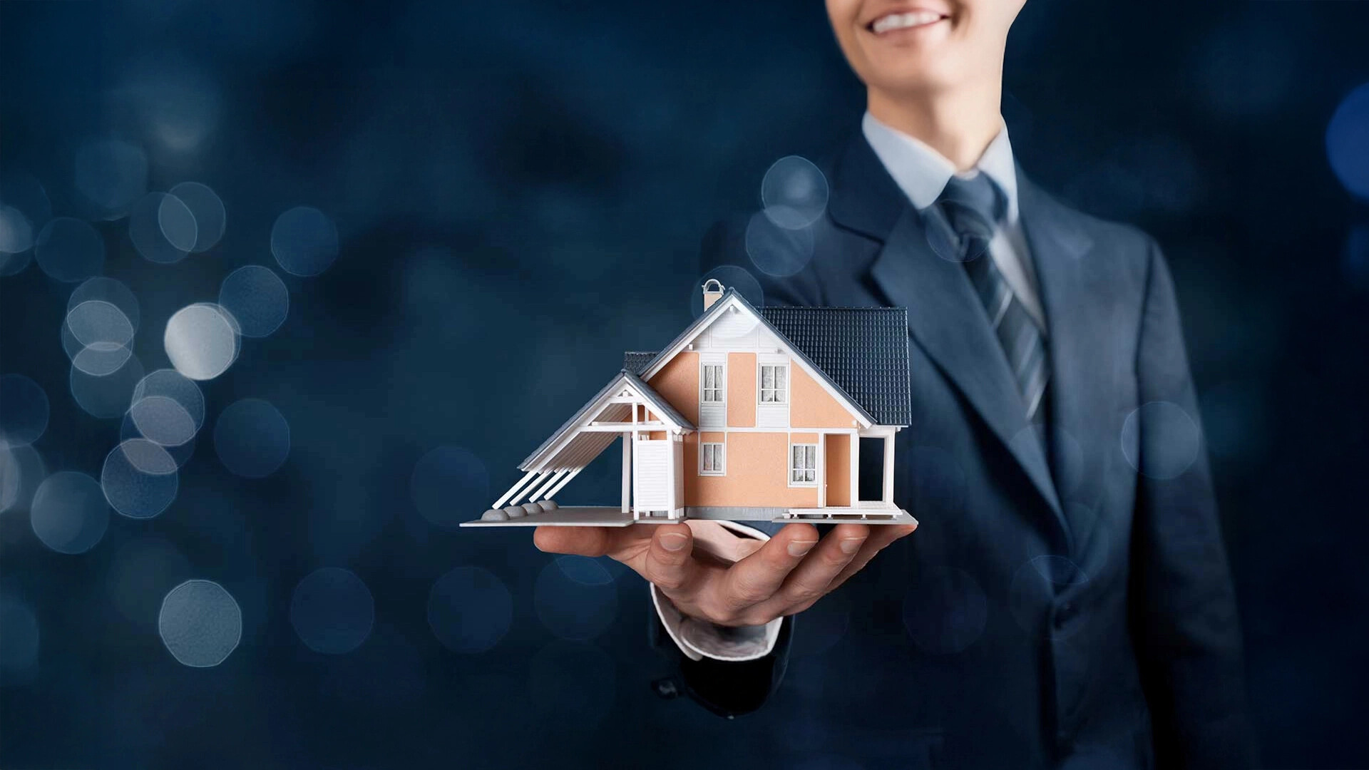 Person im Anzug hält ein Modellhaus vor dunklem Hintergrund.