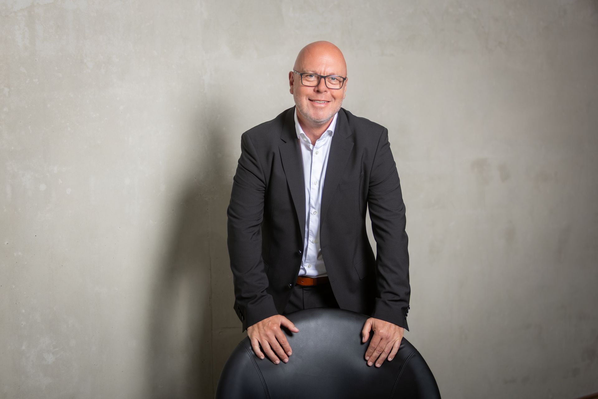 Andreas Scherer im Anzug lächelt hinter einem schwarzen Stuhl, dahinter eine graue Wand.