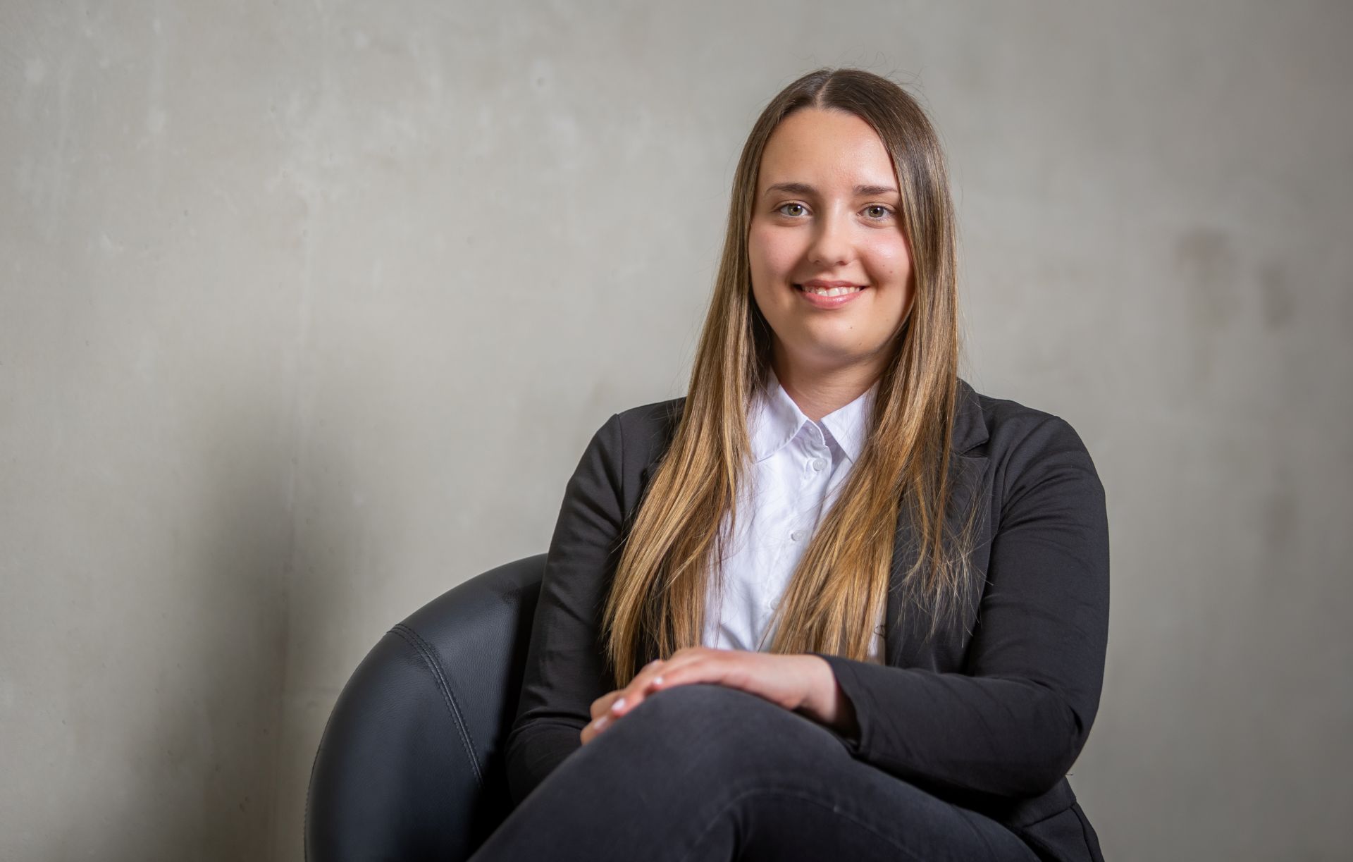 Lena Dörrschuck in Businesskleidung sitzt lächelnd auf Stuhl vor Grau.