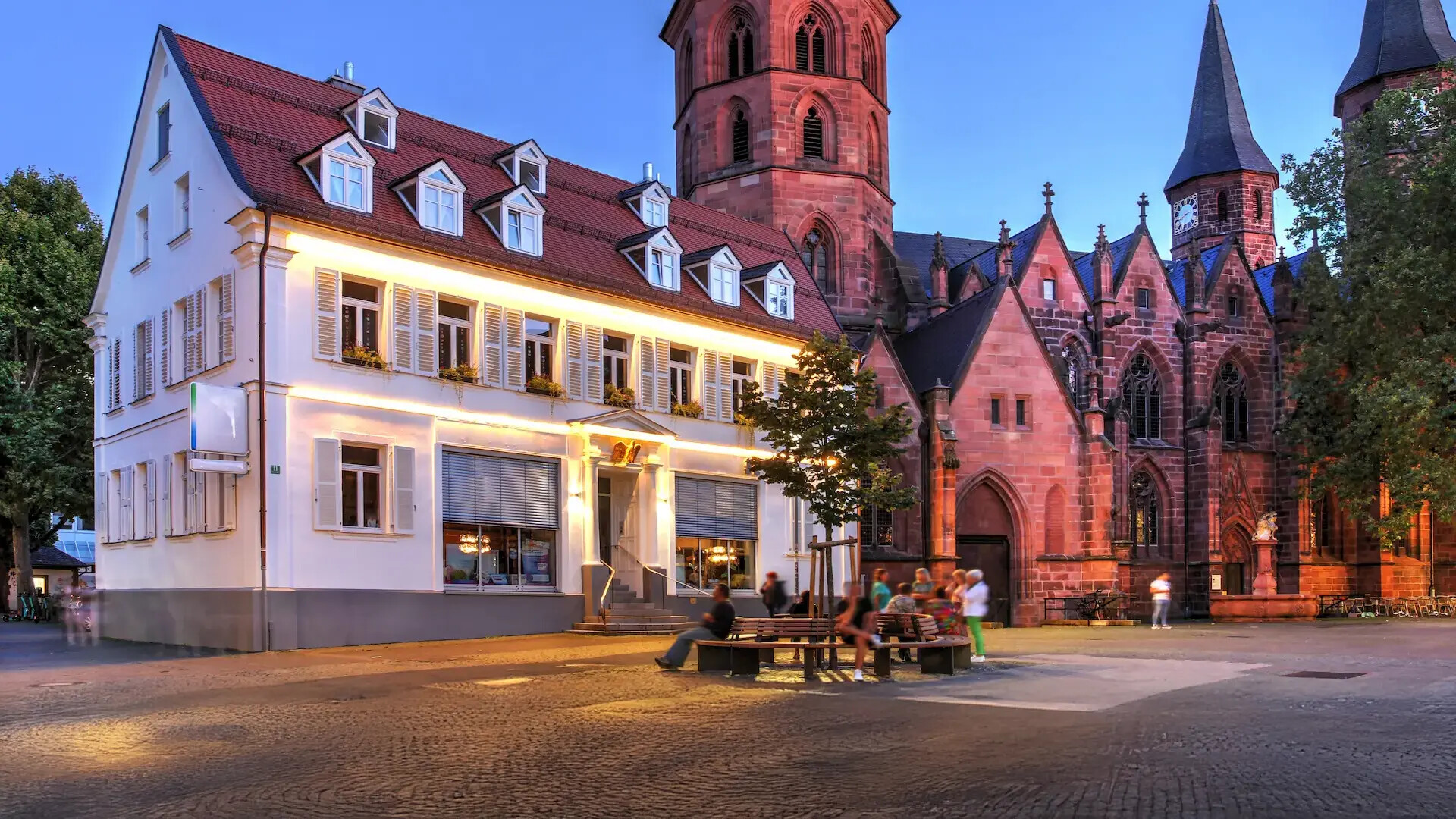 Leute sitzen bei Abenddämmerung auf Bänken vor beleuchtetem Gebäude.