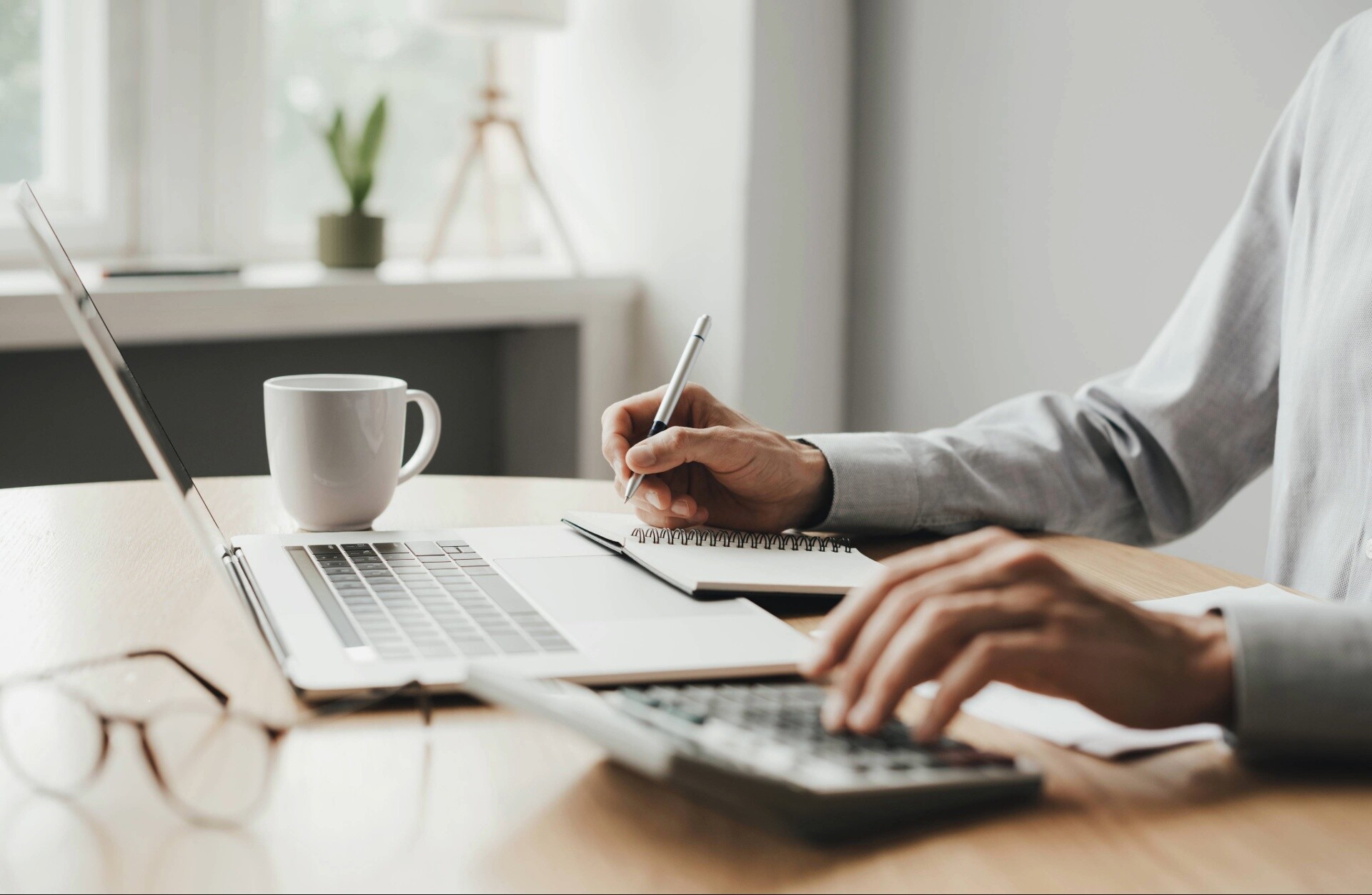 Ein Mann sitzt an einem Schreibtisch vor einem Laptop, tippt auf einem Taschenrechner und notiert etwas in einen Notizblock