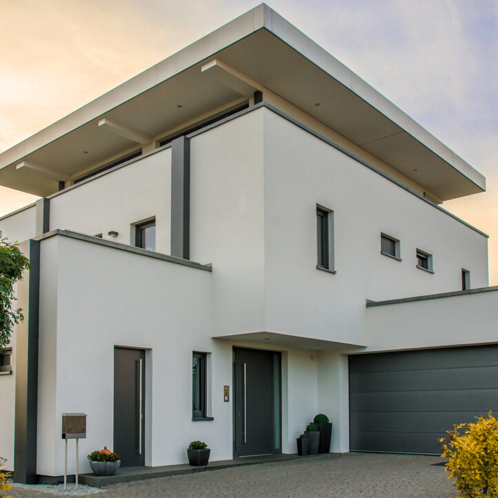 Modernes weißes zweistöckiges Haus mit Flachdach, grauem Garagentor und minimaler Bepflanzung bei Sonnenuntergang.