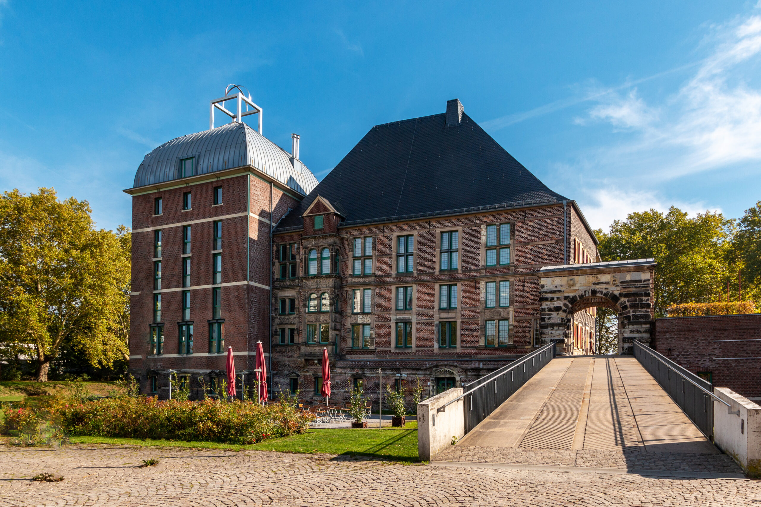 Historisches Backsteingebäude mit großen Fenstern, bogenförmigem Eingangstor und modernem Dachausbau unter blauem Himmel.