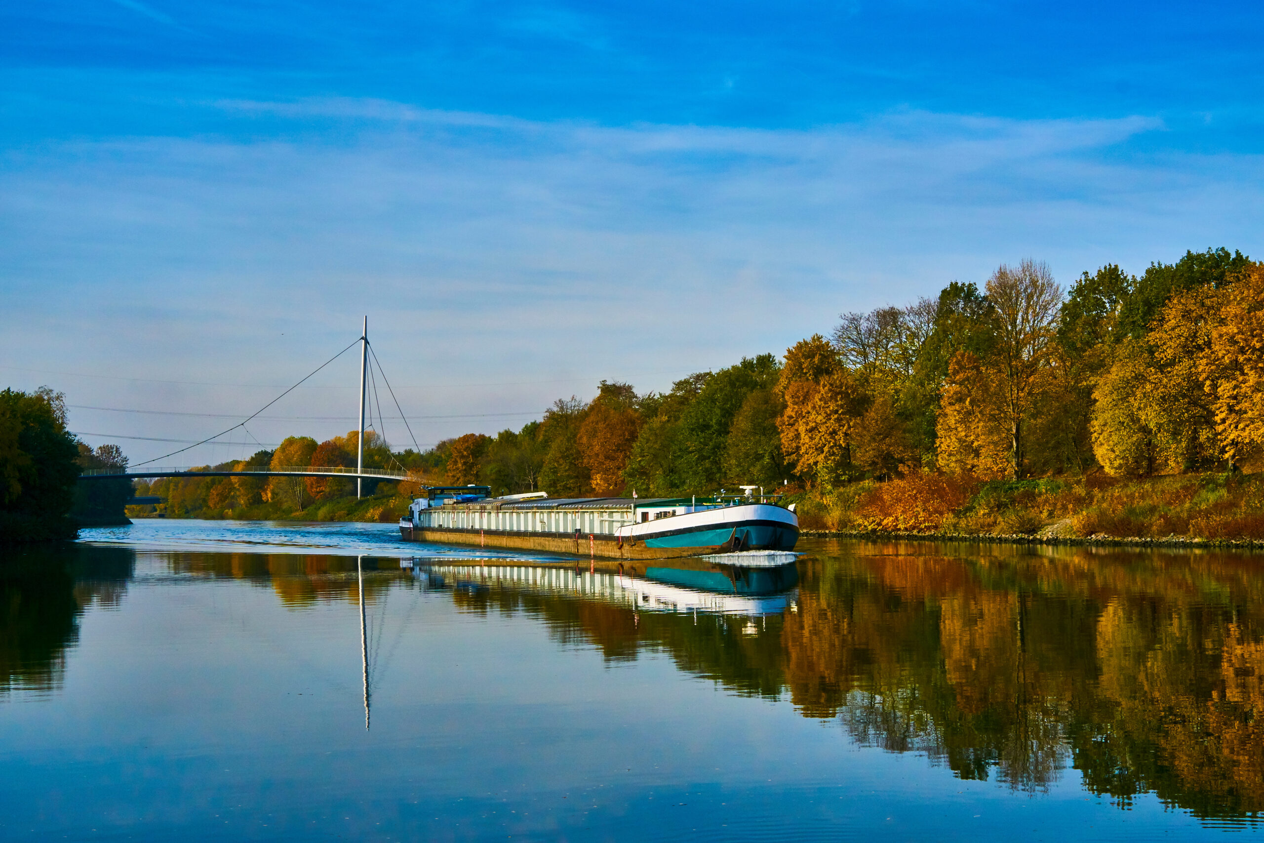Ein Lastkahn fährt auf einem ruhigen Fluss mit Herbstbäumen und einer Brücke im Hintergrund.