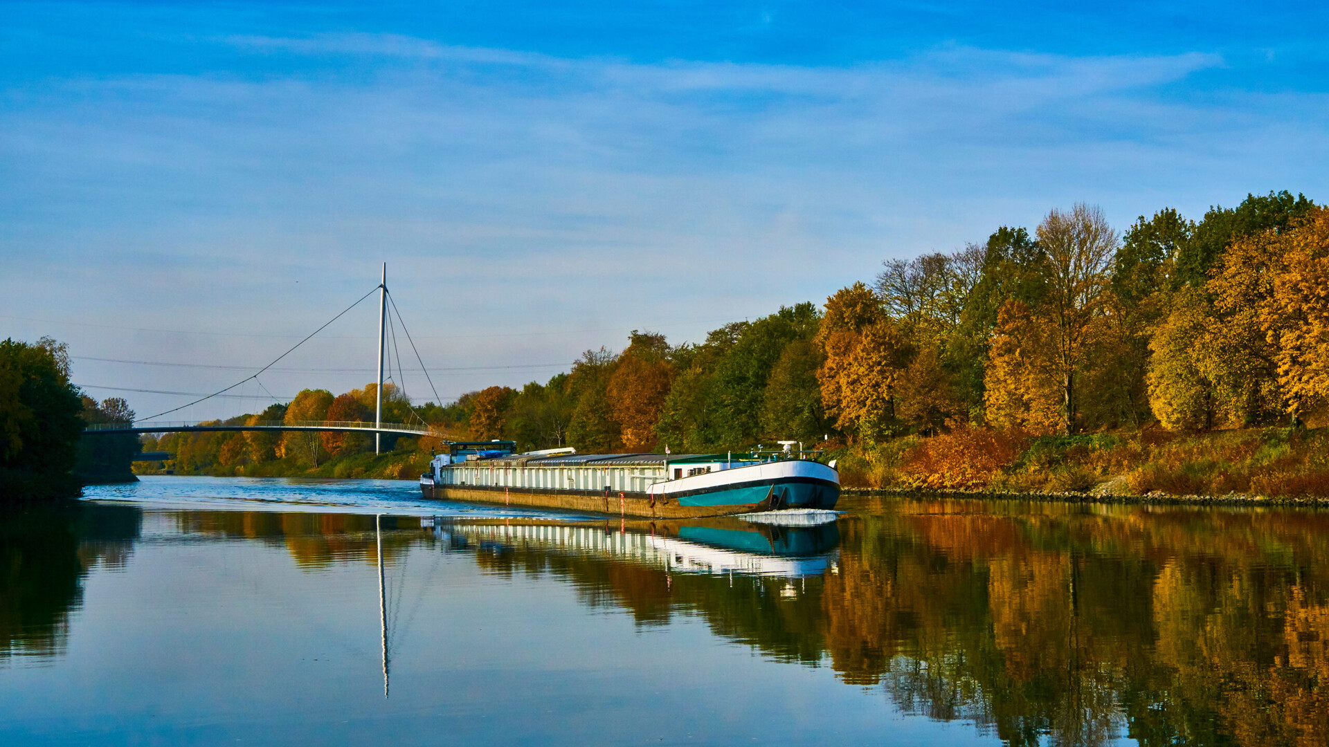 Ein Lastkahn fährt auf einem ruhigen Fluss mit Herbstbäumen und einer Brücke im Hintergrund.