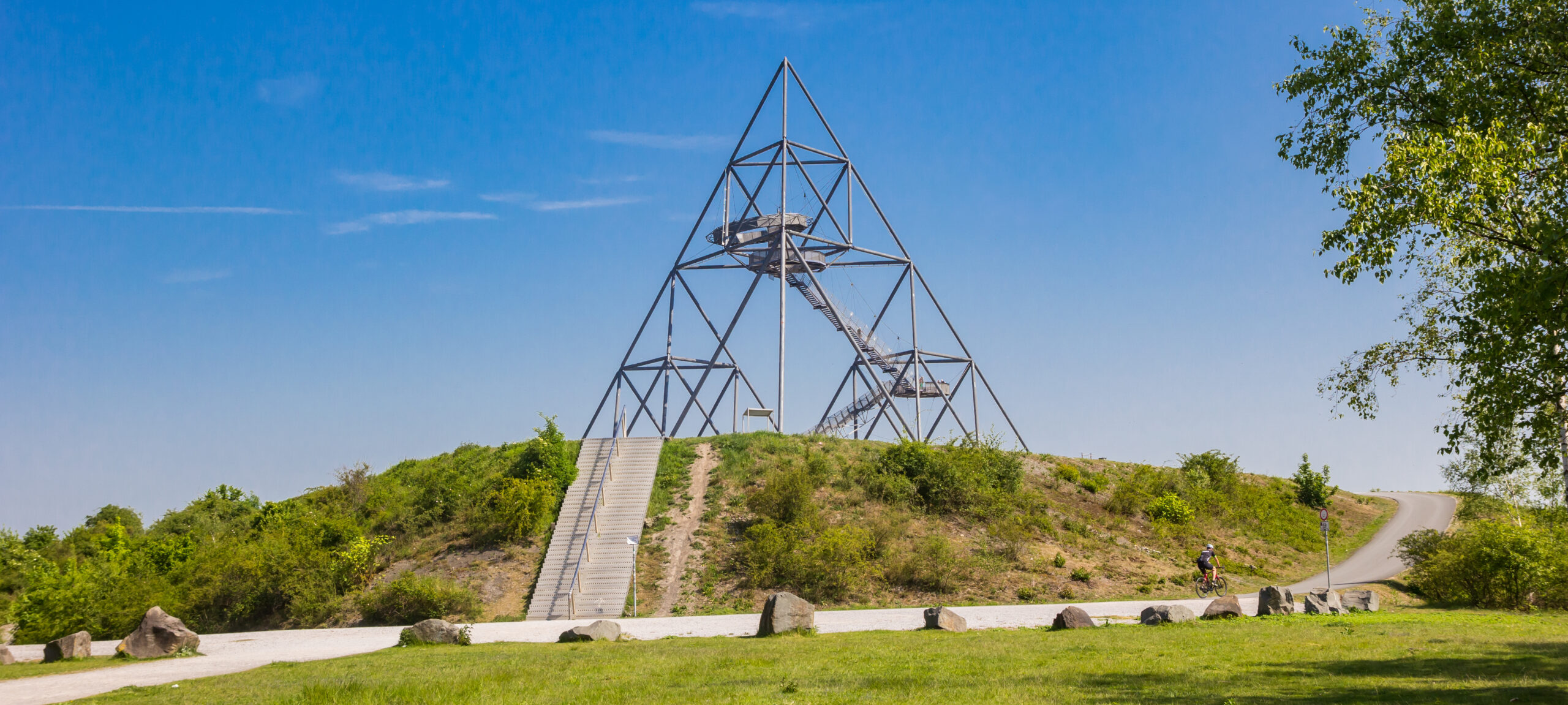 Tetraedrischer Aussichtsturm aus Metall auf einem grasbewachsenen Hügel mit Treppen, unter einem klaren blauen Himmel.