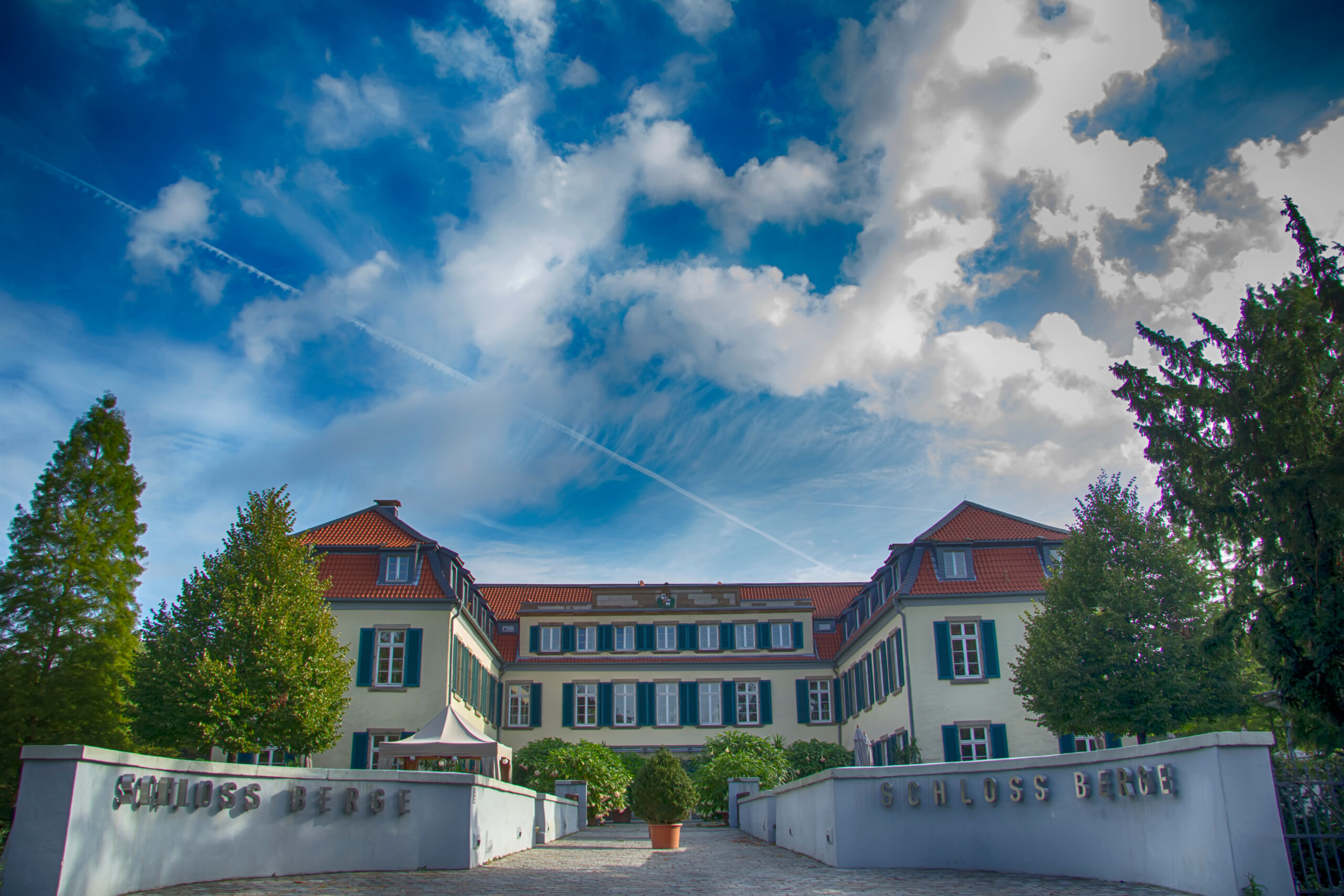 Ein großes weißes Gebäude mit grünen Fensterläden mit der Aufschrift Schloss Berge unter einem strahlend blauen Himmel mit Wolken.