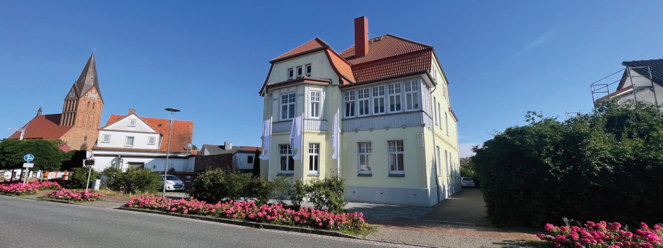 Gelbes Haus mit rotem Dach, Blumen davor, Kirche im Hintergrund.
