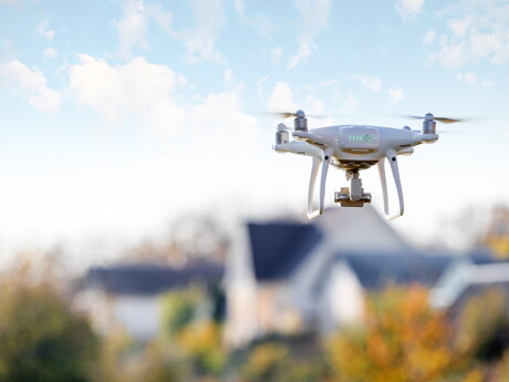 Weiße Drohne mit Kamera, die über verschwommene Häuser und Bäume fliegt.