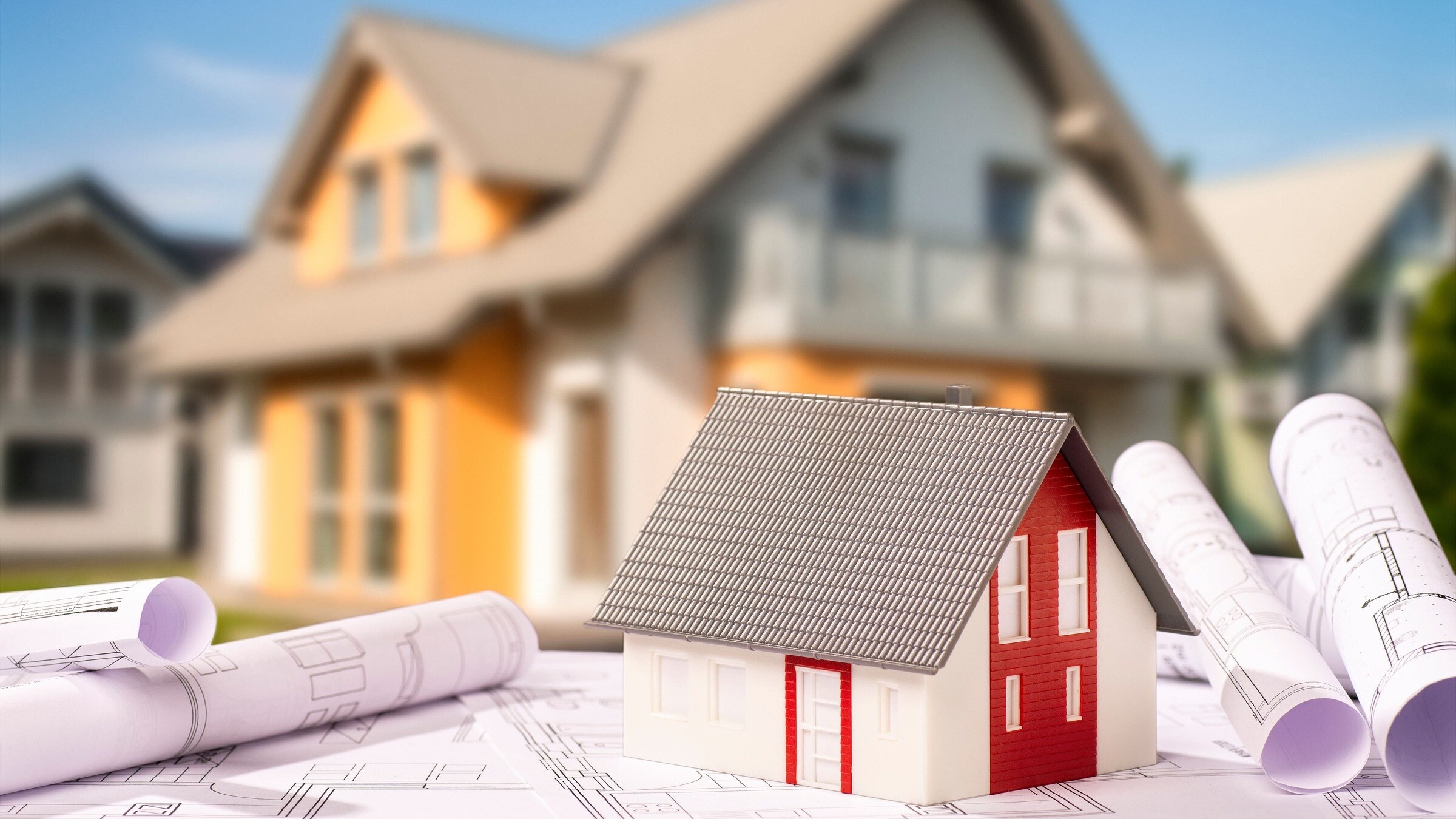 Modellhaus und Baupläne, im Hintergrund ein unscharfes Einfamilienhaus