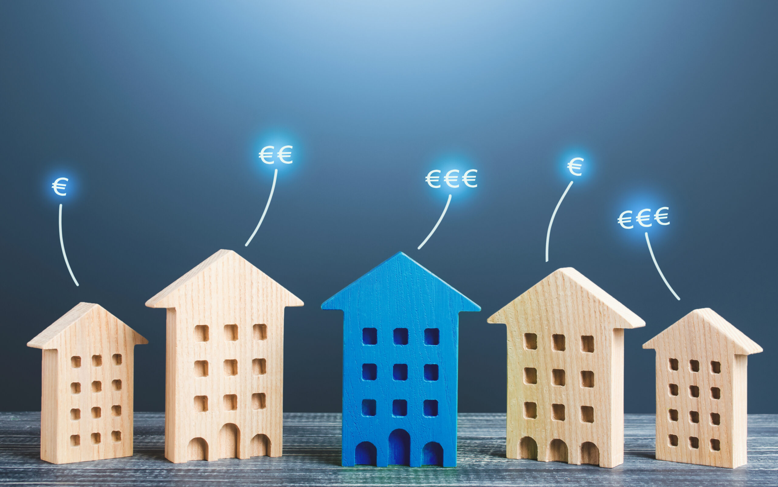 Fünf Holzhaus-Modelle, das mittlere blau, mit Euro-Symbolen.