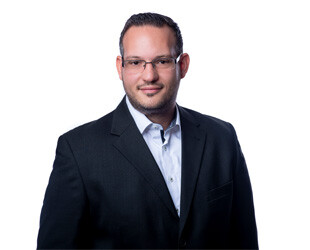 Mann in dunklem Anzug und Brille vor weißem Hintergrund.