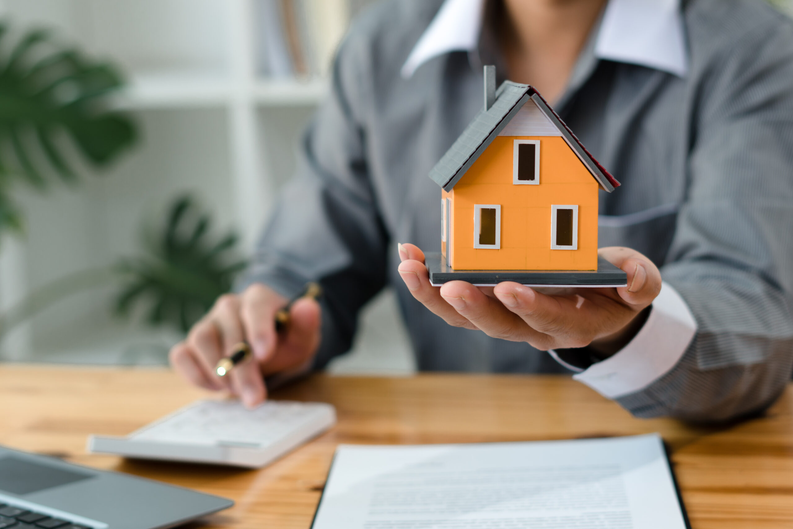 Hand hält Modellhaus, davor liegen Dokumente und Taschenrechner.