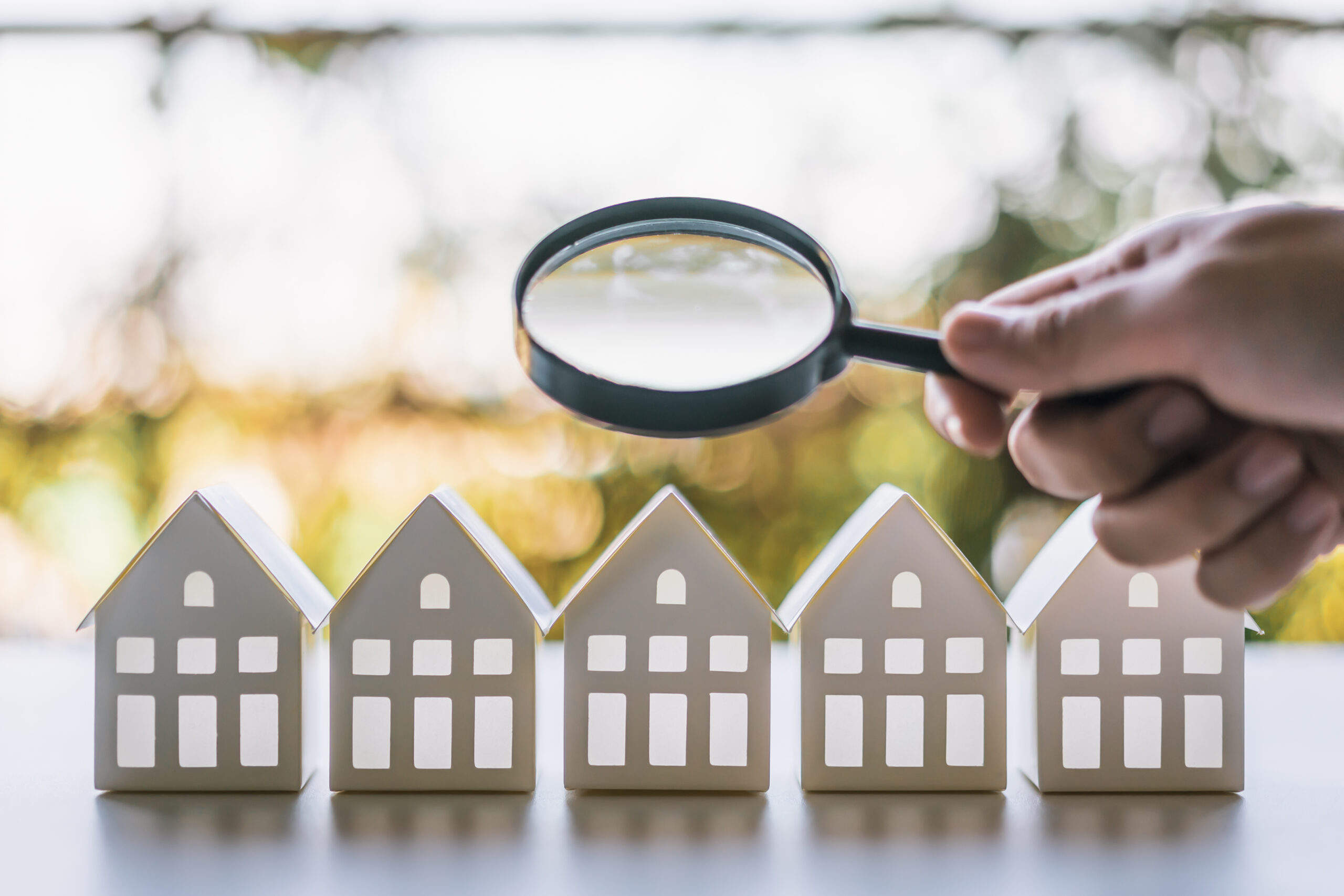 Hand mit Lupe über weißen Mini-Häusern auf einem Tisch.