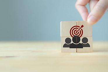 Hand setzt Holzblöcke mit Zielsymbol über drei Personen-Silhouetten.