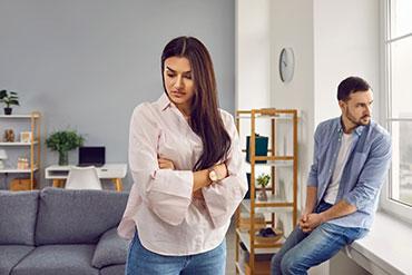 Frau mit verschränkten Armen blickt vom Mann am Fenster weg.