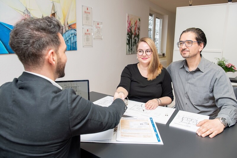 Handschlag zwischen Pärchen und Mann. Alle 3 sitzen am Tisch. Im Hintergrund sieht man Bilder aufgehängt abstrakte Kunst