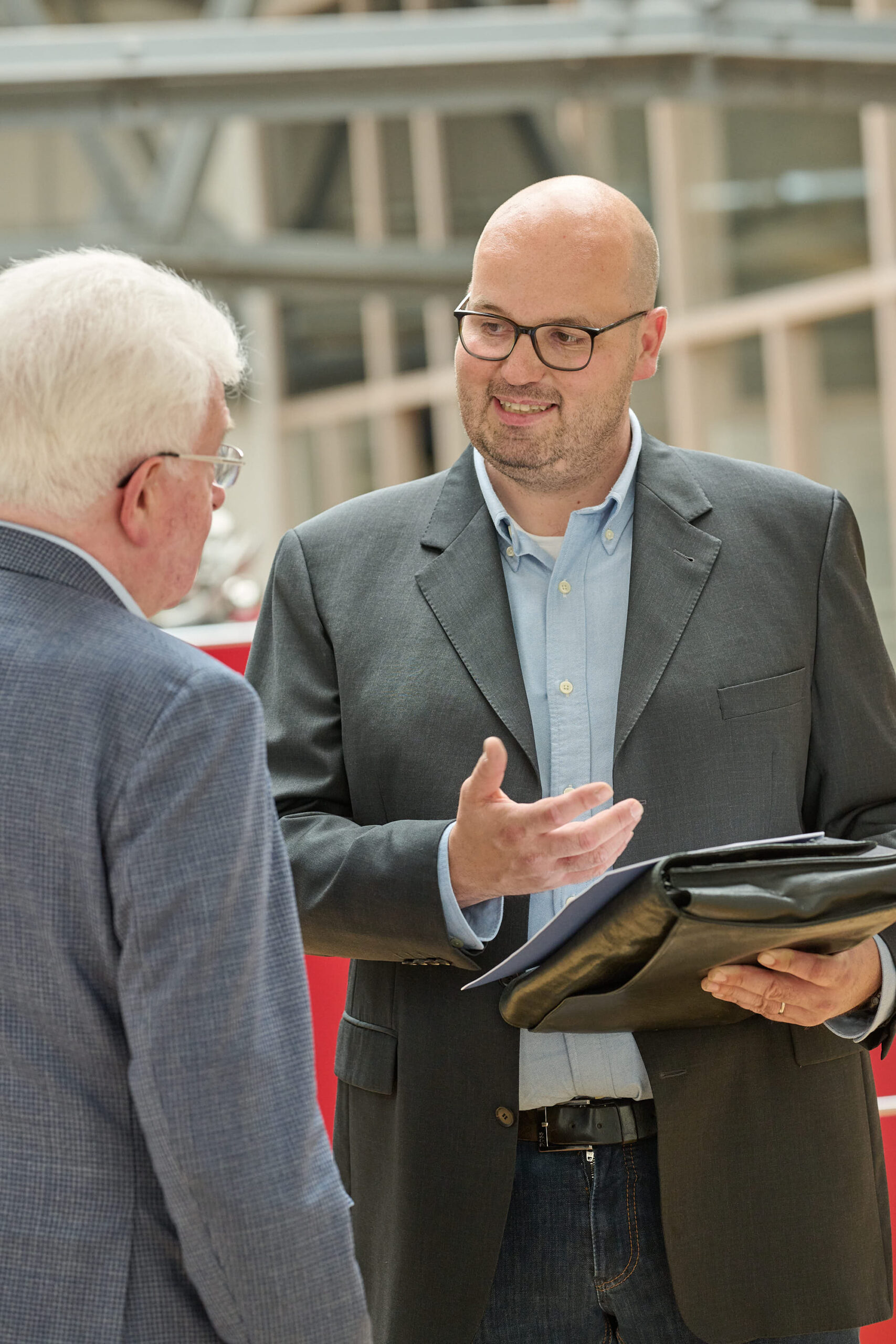 Zwei Männer im Anzug reden drinnen, einer hält eine Mappe.