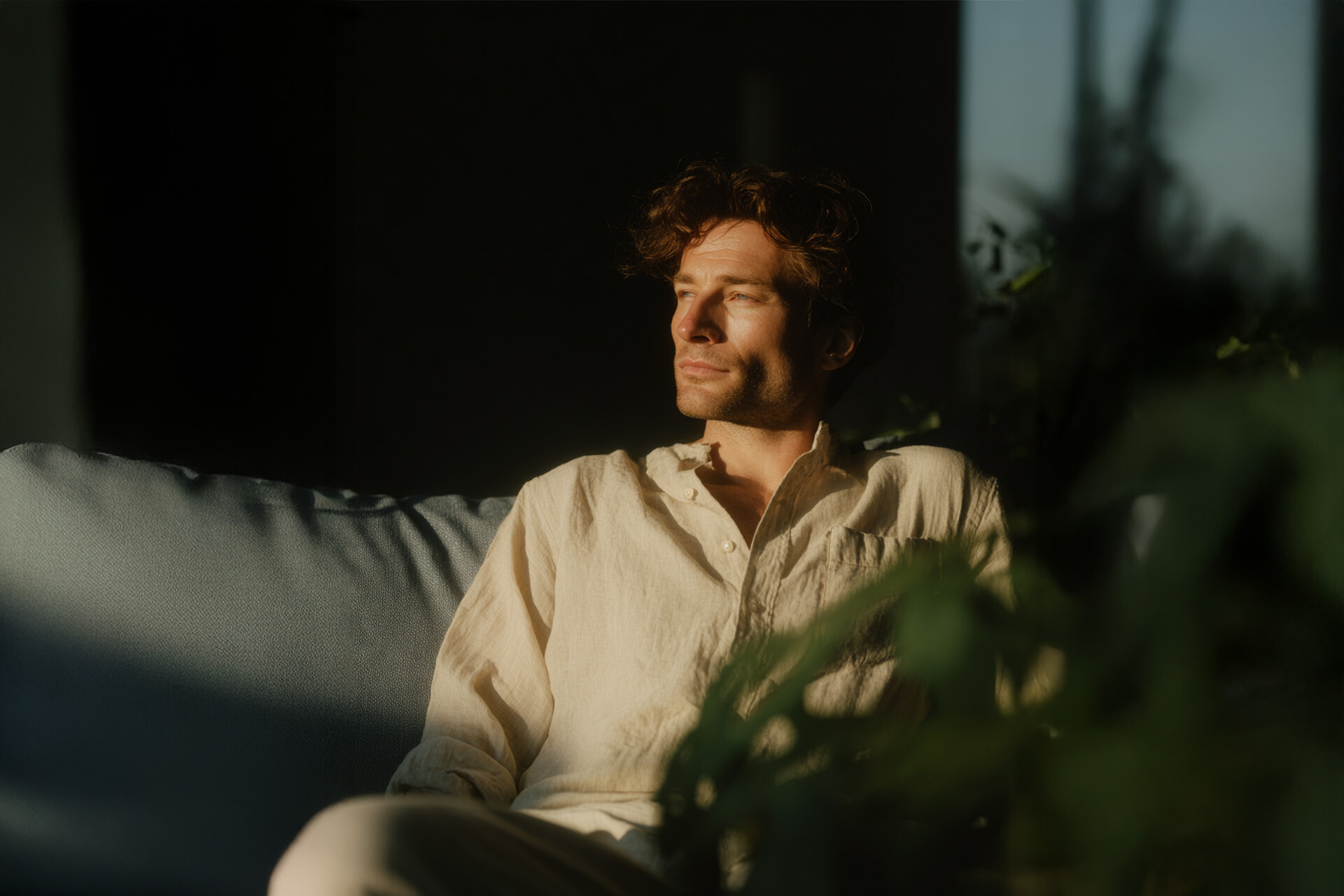 Man in beige shirt sits on sofa, sunlight, plants in front.